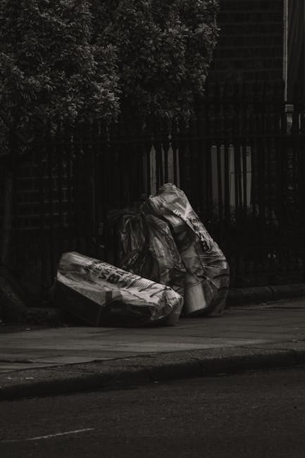 A close-up view of an outdoor rubbish pile situated on a paved surface beside a low stone wall. The heap contains black plastic rubbish bags, some partially torn open revealing mixed waste, along with