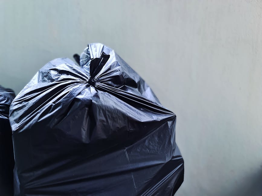 A narrow urban alleyway containing a large, black, fabric refuse sack in the foreground, positioned on uneven, dark asphalt ground with visible dirt and debris. Behind the sack, there are stacked card